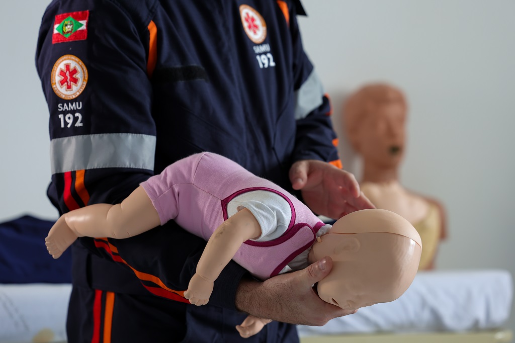 Profissional do SAMU 192, vestindo uniforme com brasão de Santa Catarina, realiza manobra de primeiros socorros em um boneco infantil. O boneco está de bruços sobre o braço do profissional, simulando atendimento a bebê engasgado. Ao fundo, há outros manequins usados para treinamentos.
