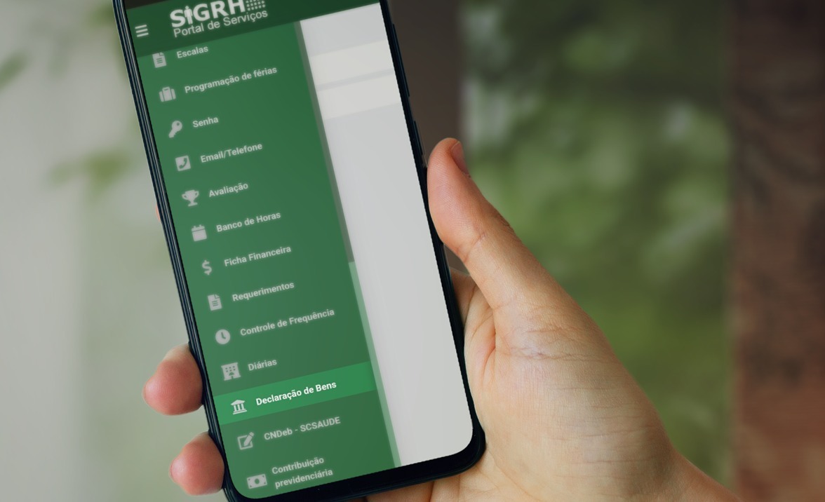 Pessoa segura um smartphone exibindo o portal SIGRH – Sistema Integrado de Gestão de Recursos Humanos – com o menu lateral visível. Entre as opções listadas estão: Escalas, Programação de férias, Senha, Email/Telefone, Avaliação, Banco de Horas, Ficha Financeira, Requerimentos, Controle de Frequência, Diárias e, em destaque, "Declaração de Bens".