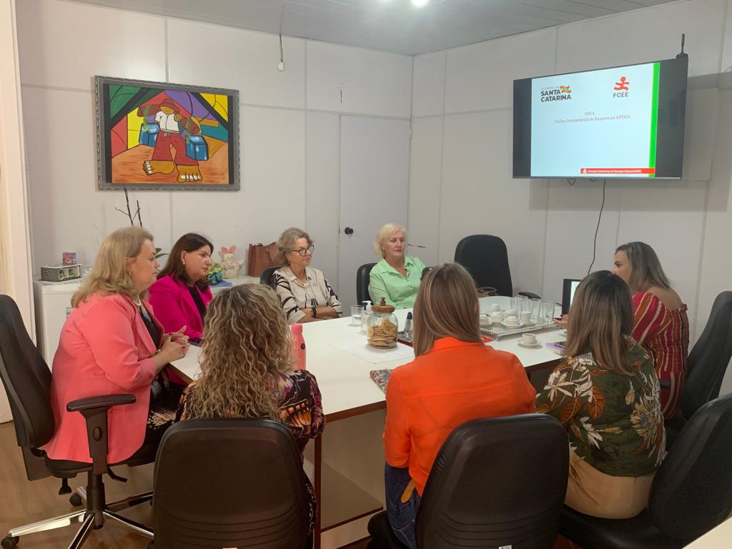 Reunião de oito mulheres sentadas ao redor de uma mesa retangular branca em uma sala de reuniões. Elas estão envolvidas em uma conversa, com copos, xícaras, cadernos e um lanche sobre a mesa. Na parede ao fundo, há uma televisão exibindo o logotipo do Governo de Santa Catarina e da FCEE com o título "Núcleo Intersetorial de Suporte ao APOIA". Também há um quadro colorido com uma pintura de estilo moderno.