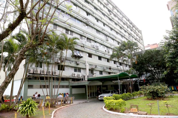 Fachada de um prédio hospitalar de vários andares, com muitas janelas equipadas com aparelhos de ar-condicionado. Na entrada principal, há uma marquise verde e um carro estacionado. À frente do prédio, há um jardim com árvores, arbustos e pessoas caminhando. O ambiente transmite uma aparência institucional e acolhedora.