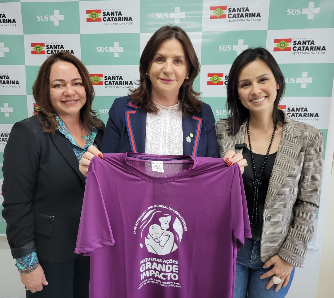 Três mulheres sorridentes posam segurando uma camiseta roxa com a frase “Pequenas ações, grande impacto” e a ilustração de uma mãe com um bebê no colo. Ao fundo, painel com logomarcas do Governo de Santa Catarina, Secretaria da Saúde e SUS. A imagem celebra o Dia Mundial da Prematuridade.