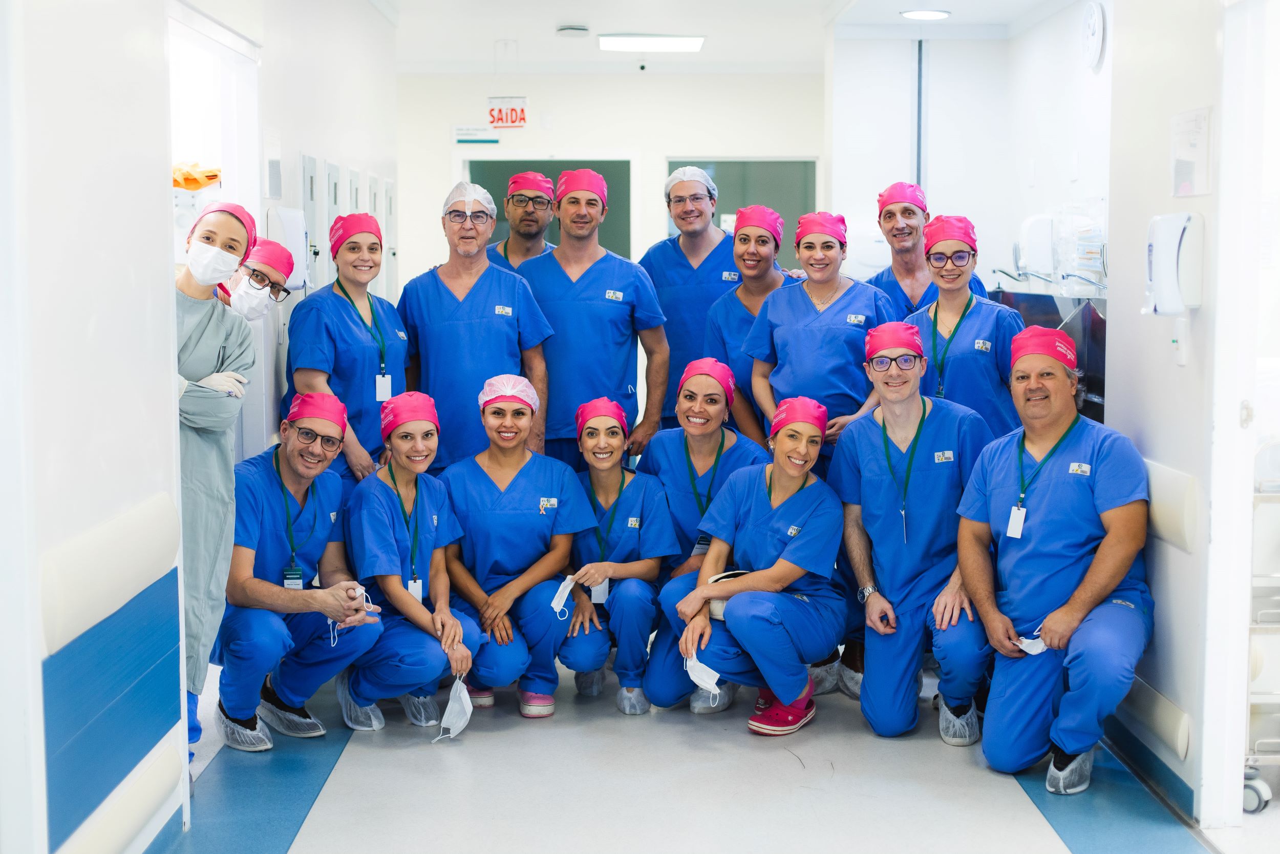 Grupo de profissionais de saúde usando uniforme azul e toucas rosas, posando juntos em corredor de hospital, sorrindo para a câmera.