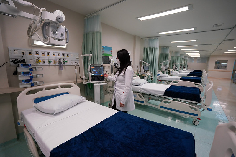 Profissional da saúde em pé ao lado de um leito hospitalar moderno, ajustando um monitor de sinais vitais. A sala é ampla, iluminada e equipada com diversas camas organizadas, todas com lençóis brancos e cobertores azuis. Equipamentos médicos estão fixados nas paredes ao lado dos leitos, prontos para uso.