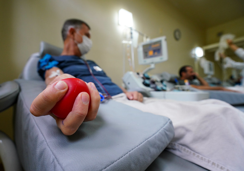 Homem doa sangue em hemocentro. Ele está deitado em uma cadeira própria para coleta, usando máscara facial, enquanto aperta uma bola vermelha com a mão para estimular a circulação. Ao fundo, outros doadores também participam da ação solidária. A imagem representa a importância da doação voluntária de sangue.