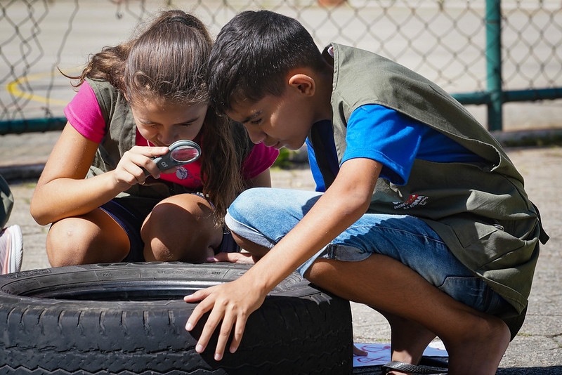 Criança menina e menino, usando coletes verdes, observam atentamente um pneu com uma lupa em um ambiente externo ensolarado. A atividade parece educativa, com foco em investigação ou ciência, e ocorre próximo a uma cerca de arame.