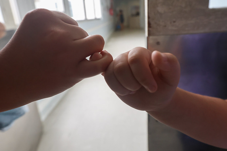 Duas crianças fazem um gesto de 'pacto do mindinho', entrelaçando os dedos mínimos em um corredor iluminado por luz natural. A imagem transmite sensações de amizade, confiança e afeto infantil.