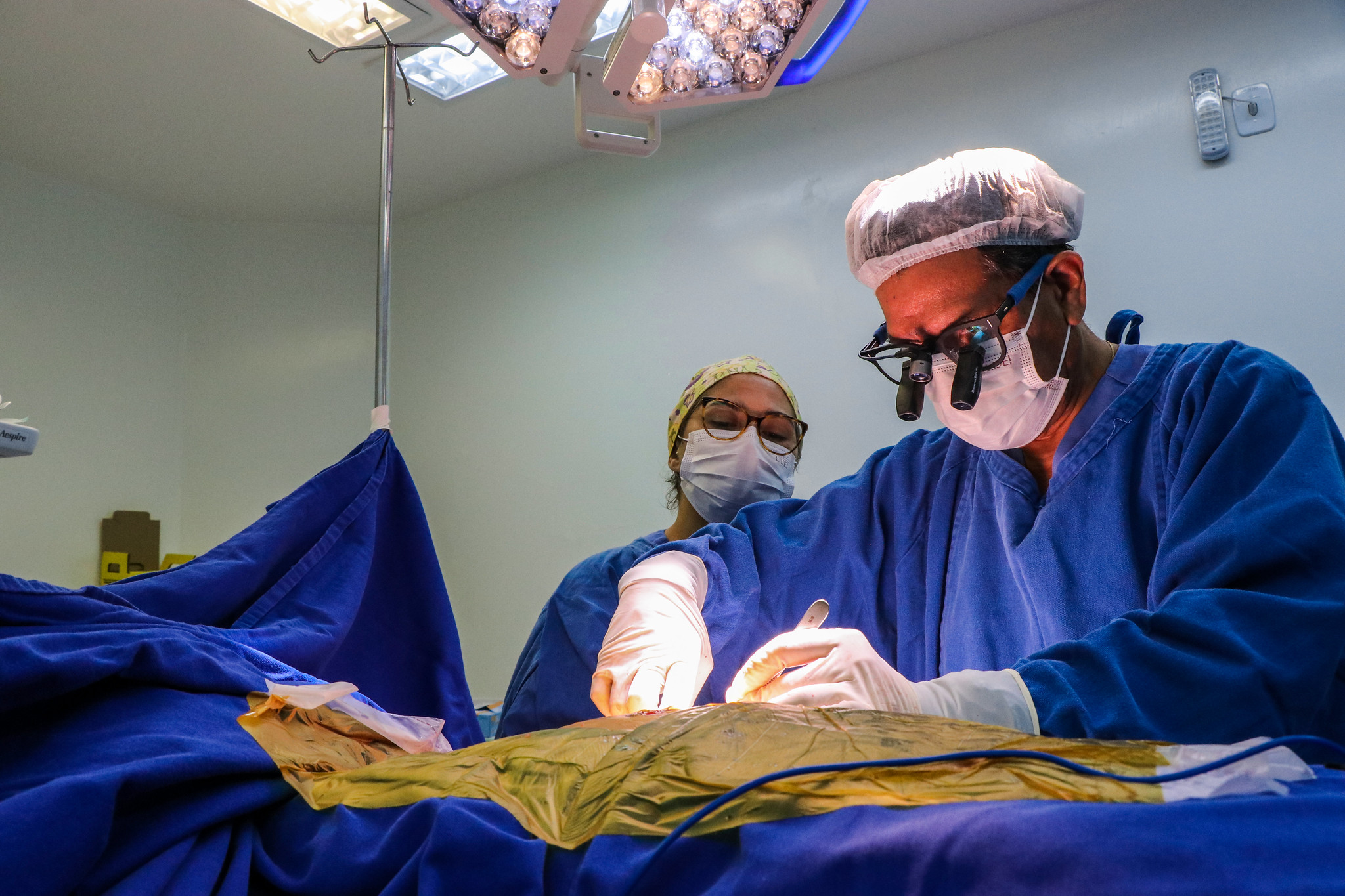 Cirurgiões vestidos com roupas hospitalares azuis realizam um procedimento em centro cirúrgico. Um dos profissionais, usando lupa cirúrgica e touca, opera com precisão sob luzes intensas do teto, enquanto outro auxilia ao fundo, ambos com máscaras e luvas.