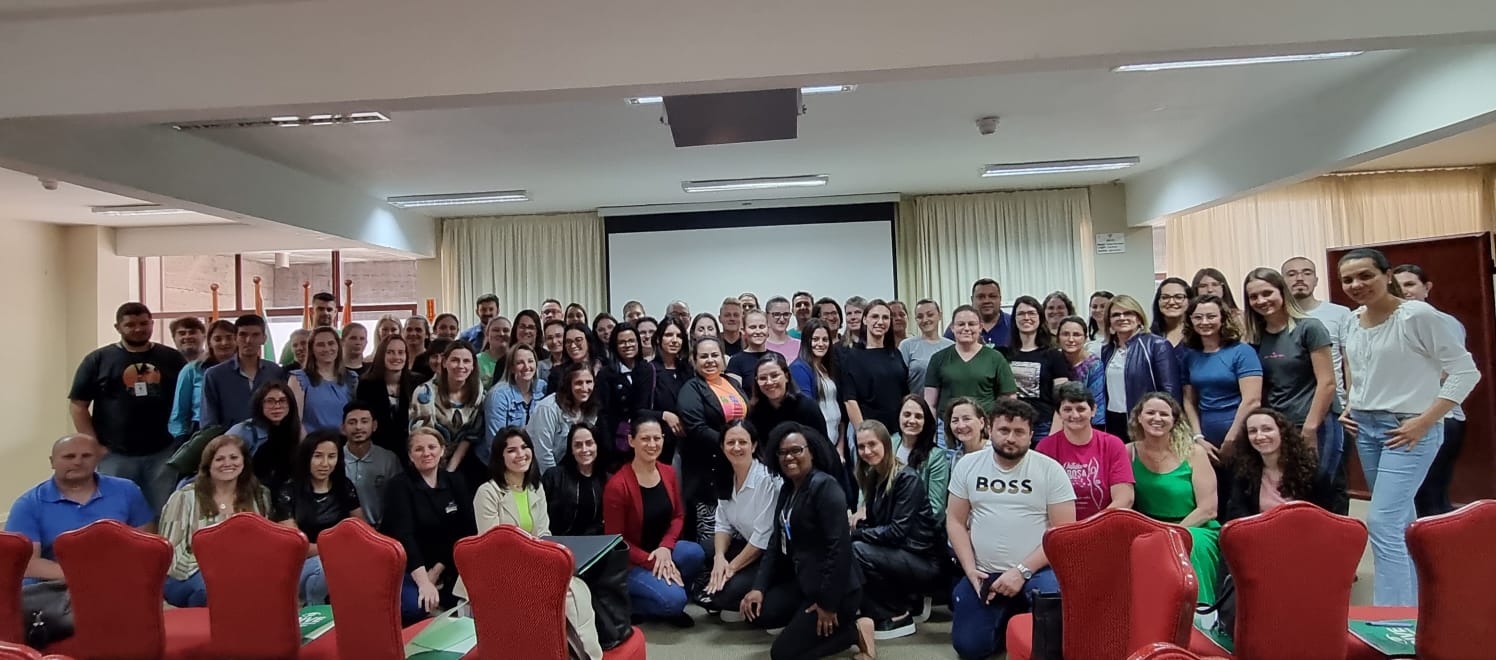 Grande grupo de pessoas posando para foto em um auditório com cadeiras vermelhas e tela de projeção ao fundo, durante um evento ou treinamento.