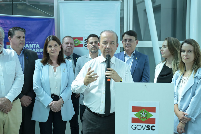 Autoridade do Governo de Santa Catarina faz discurso ao microfone, cercado por equipe de governo, em cerimônia oficial com banners do GOVSC ao fundo.