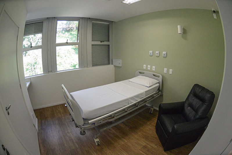 Quarto hospitalar individual com cama ajustável ao centro, poltrona preta ao lado e parede verde clara com tomadas e conexões médicas. Janela ampla ao fundo com vista para árvores.