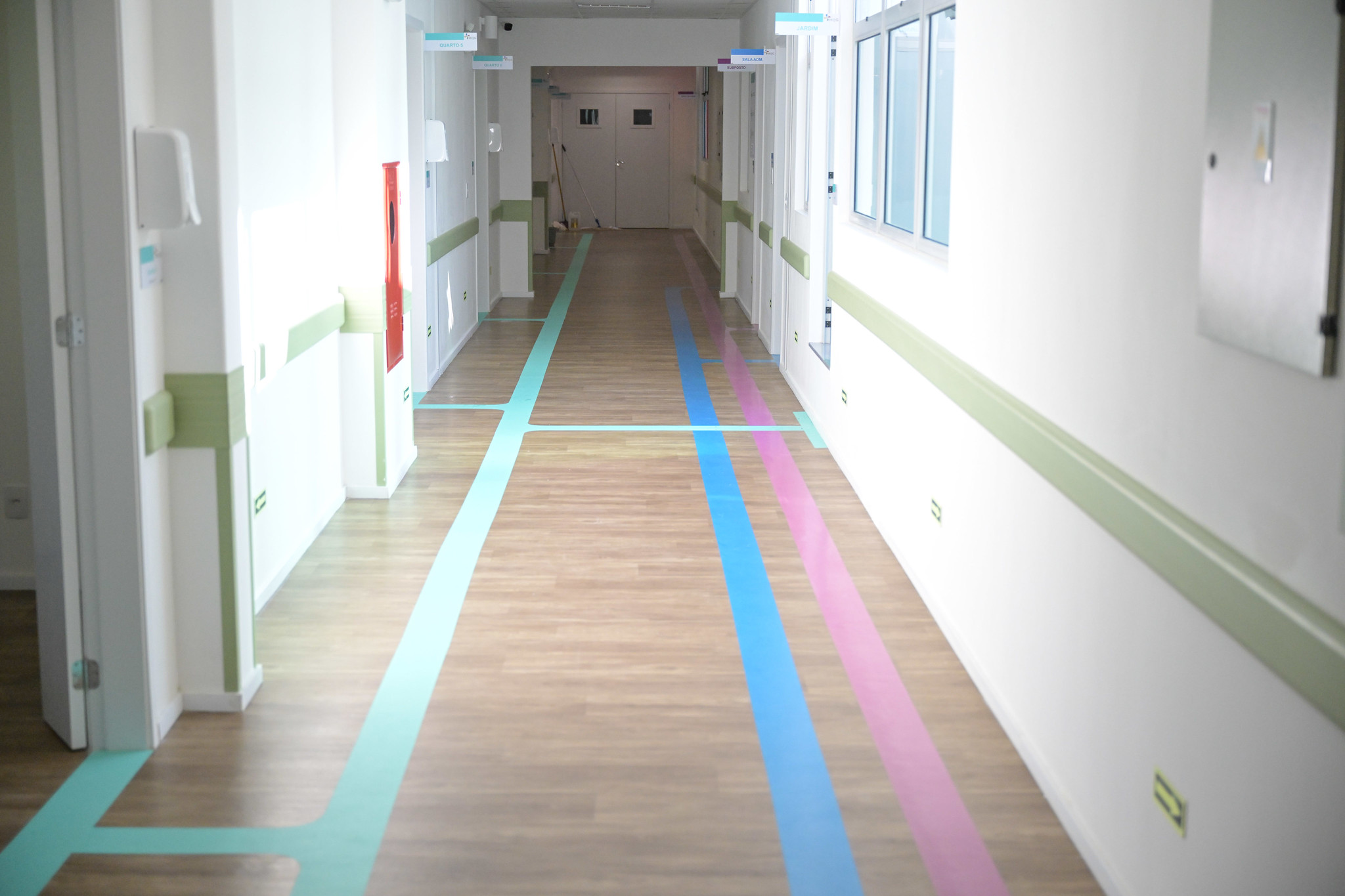 Corredor interno de hospital com piso de madeira e faixas coloridas no chão indicando caminhos para diferentes setores. Paredes brancas com detalhes verdes e iluminação natural.