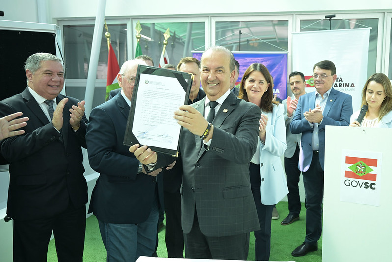 Autoridade do Governo de Santa Catarina exibe documento oficial assinado, rodeado por outras autoridades e profissionais, em cerimônia com logotipo do Governo do Estado ao fundo.