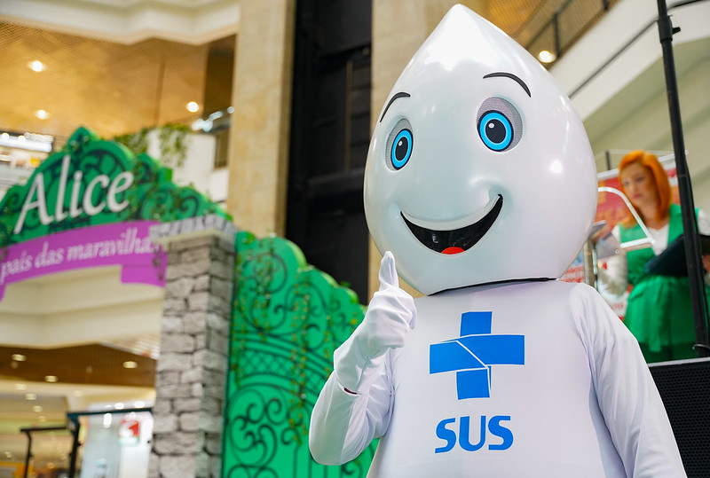 Mascote Zé Gotinha, símbolo das campanhas de vacinação do SUS, fazendo sinal de positivo em ambiente decorado com tema de Alice no País das Maravilhas.