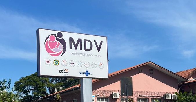 Placa da Maternidade Darcy Vargas (MDV), com logotipo estilizado de uma figura adulta segurando uma criança em rosa e preto. Abaixo do nome constam os logotipos do Governo de Santa Catarina, do SUS e de outras entidades de saúde. Ao fundo, prédio de cor rosa e céu azul claro.