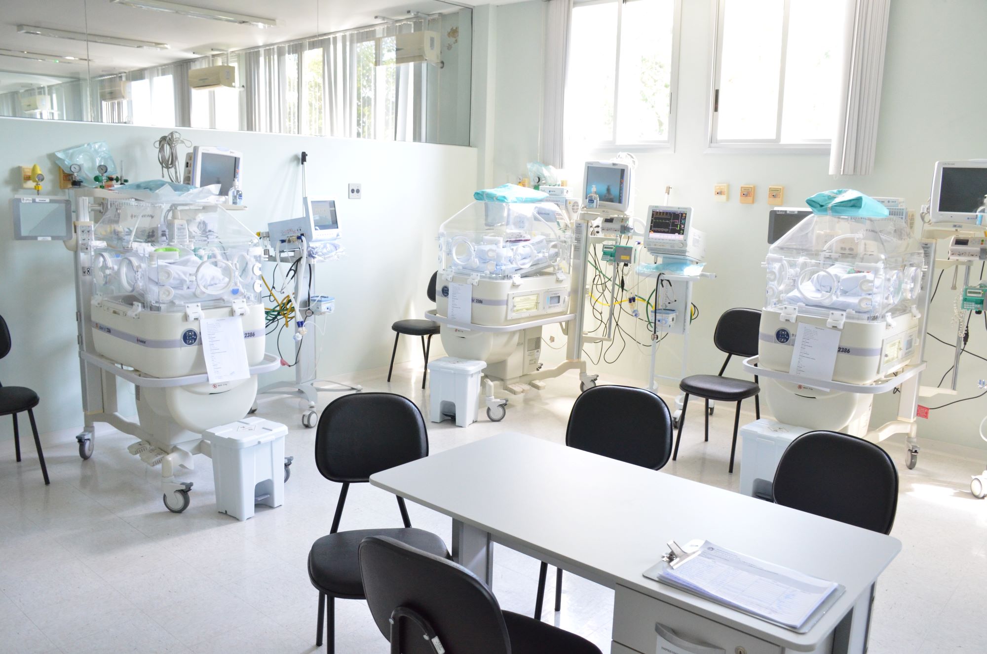 Sala de unidade neonatal com várias incubadoras modernas, monitores multiparâmetros e equipamentos médicos ao redor. Há cadeiras e uma mesa com prancheta no centro, sugerindo um espaço preparado para atendimento e acompanhamento de recém-nascidos em estado delicado.