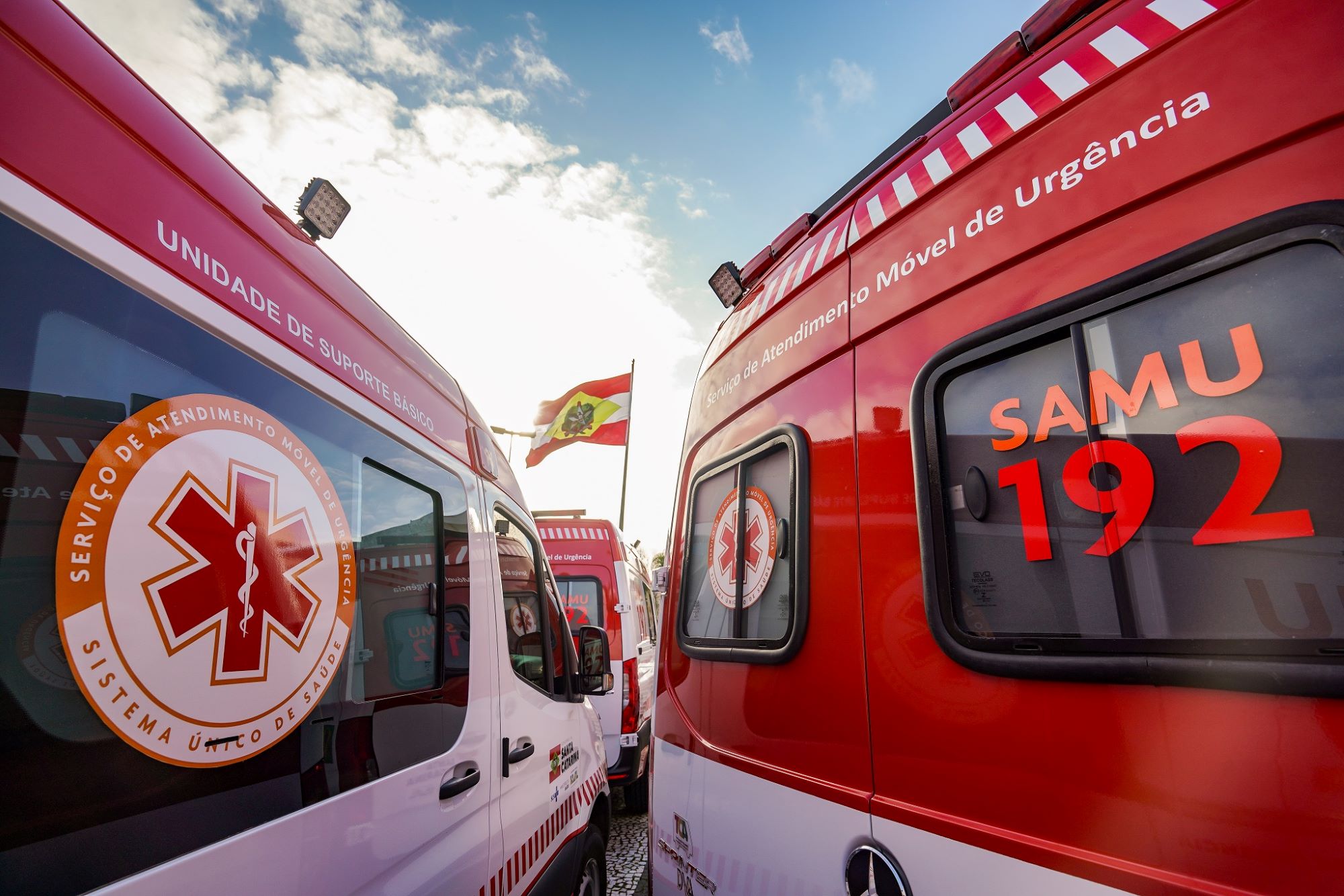 A imagem mostra ambulâncias do SAMU (Serviço de Atendimento Móvel de Urgência), com destaque para a inscrição SAMU 192, que é o número de telefone de emergência no Brasil. No canto esquerdo, lê-se Unidade de Suporte Básico, e ao fundo há uma bandeira do estado de Santa Catarina, Brasil.
