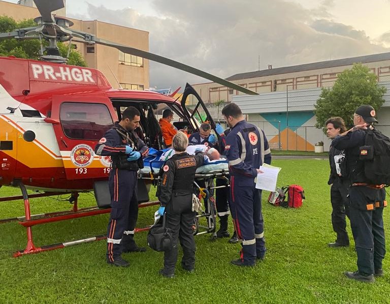   O ChatGPT disse: A imagem mostra uma equipe de atendimento de emergência realizando um transporte aeromédico. O helicóptero, identificado pelo prefixo PR-HGR e pelas marcas do Bombeiro Militar e do SAMU 192, está no solo com a porta aberta.  Vários profissionais — incluindo paramédicos, médicos e socorristas — cercam um paciente deitado em uma maca, aparentemente recebendo cuidados médicos antes ou durante o embarque/desembarque. O cenário é um gramado próximo a prédios, sugerindo que o pouso foi feito em área urbana, possivelmente em um hospital.