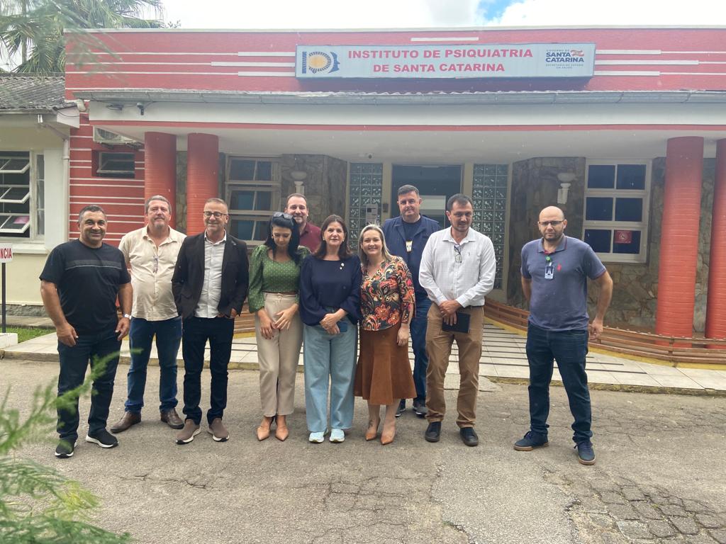 Grupo de 12 pessoas posa para foto em frente ao prédio do Instituto de Psiquiatria de Santa Catarina, com fachada vermelha e letreiro visível. A maioria sorri e a cena sugere uma visita institucional ou encontro oficial.