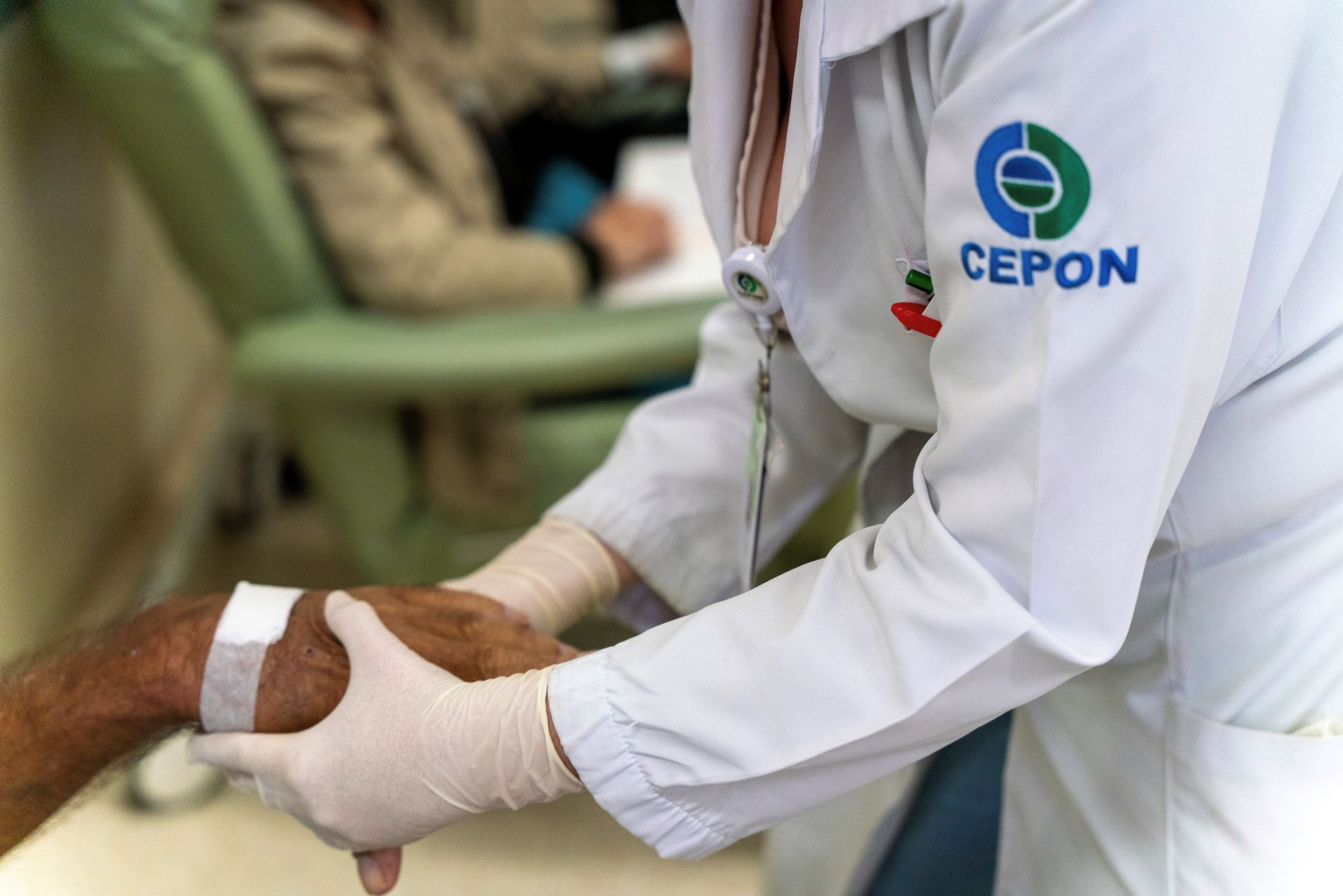 A imagem mostra um profissional de saúde vestindo jaleco branco com o logotipo do CEPON e usando luvas descartáveis, segurando com cuidado a mão de um paciente. A mão do paciente apresenta um curativo branco no dorso. Ao fundo, desfocado, há outra pessoa sentada em uma cadeira de atendimento. A cena transmite acolhimento e cuidado no ambiente hospitalar.