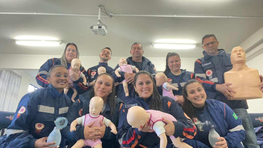 A imagem mostra um grupo de profissionais do SAMU vestindo uniformes azul-marinho com detalhes laranja e o emblema de emergência médica no peito. Eles estão em uma sala de treinamento e seguram manequins utilizados para práticas de primeiros socorros — alguns simulando bebês e outros simulando o tronco de um adulto. Dois participantes também seguram ressuscitadores manuais (Ambu bag), usados para ventilação artificial. O clima é de aprendizado e cooperação, evidenciando um treinamento de suporte básico de vida voltado para diferentes faixas etárias.