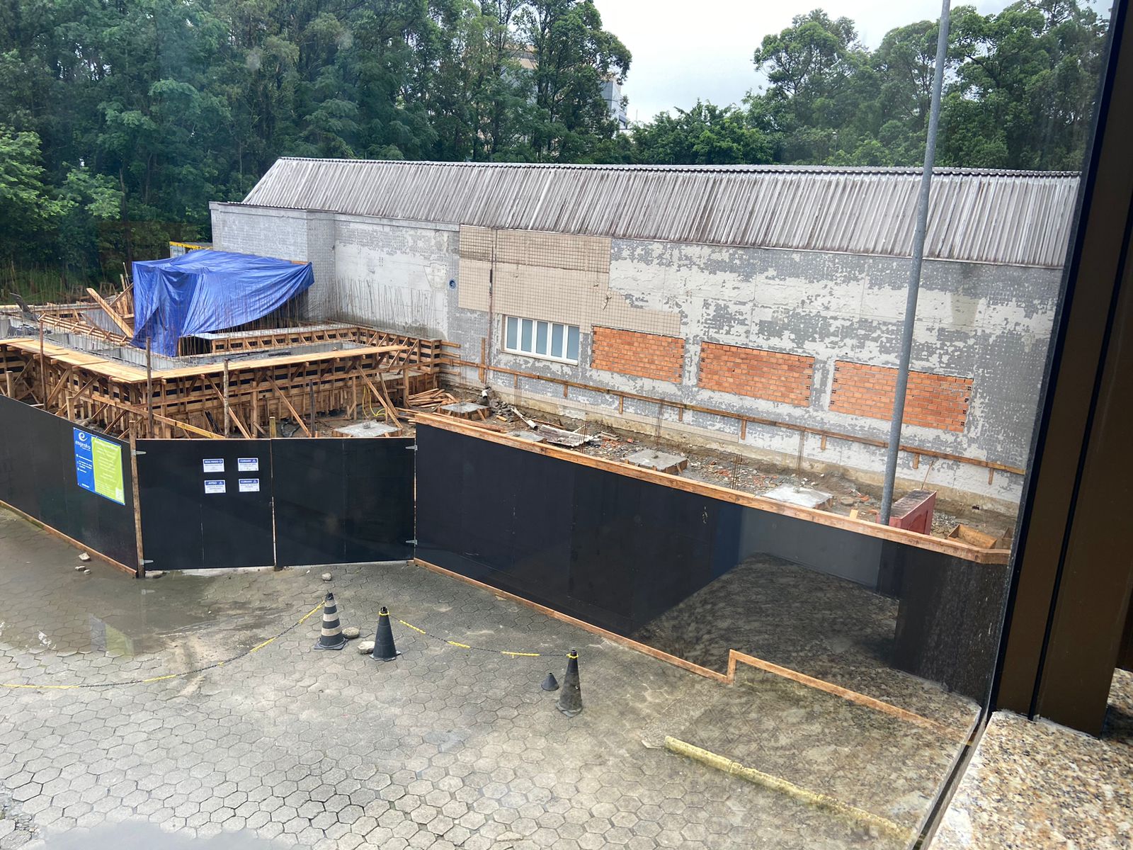 A imagem mostra um canteiro de obras visto de um ponto elevado, possivelmente de dentro de um prédio. Na área em construção, há uma estrutura de madeira sendo utilizada como forma para concreto, parcialmente coberta por uma lona azul. O prédio ao fundo tem uma fachada desgastada, com áreas de tijolo exposto e janelas. Um tapume preto com placas informativas e avisos cerca o local da obra. Na parte externa, sobre o piso de blocos intertravados, há cones de sinalização e uma faixa amarela delimitando o acesso. Ao fundo, há uma área com árvores densas, sugerindo que o local fica próximo a uma região arborizada.