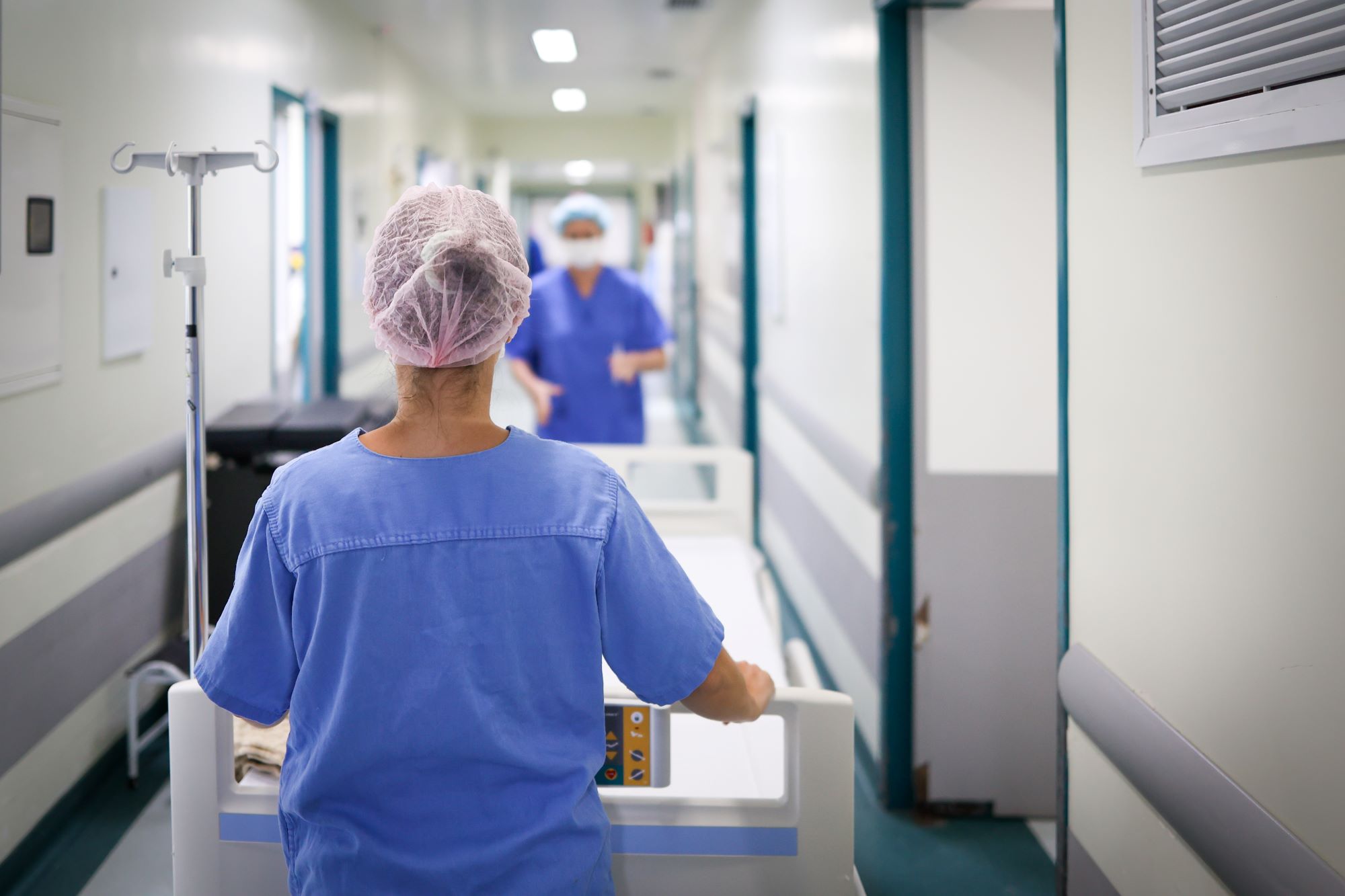 Profissional de saúde visto de costas empurrando uma maca por um corredor de hospital. A pessoa veste uniforme azul e touca descartável rosa. À frente, outro profissional também uniformizado aparece desfocado. O corredor é bem iluminado, com paredes claras e portas hospitalares.