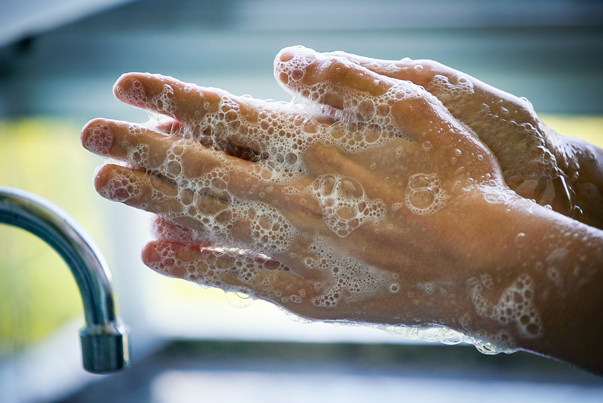Mãos sendo lavadas com sabão, cobertas de espuma, em frente a uma torneira metálica. A imagem destaca a importância da higiene das mãos para a prevenção de doenças.