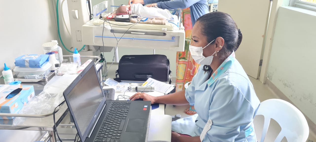 Profissional da saúde usando jaleco azul e máscara facial trabalha em um notebook dentro de uma unidade neonatal. Ao fundo, um bebê recém-nascido está deitado em uma incubadora, sendo cuidado por outra pessoa. A sala tem equipamentos médicos, materiais de enfermagem e iluminação natural vinda da janela.