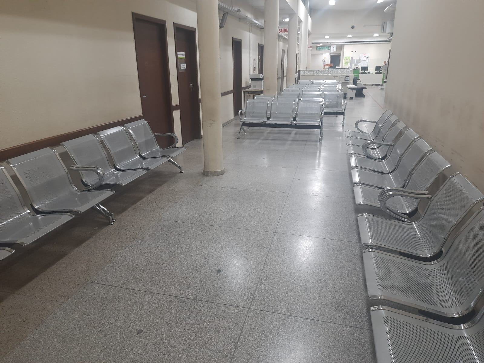 A imagem mostra uma sala de espera interna, provavelmente de um hospital, clínica ou repartição pública. Há fileiras de cadeiras metálicas com acabamento prateado dispostas dos dois lados do corredor, todas vazias.  O chão é de granito claro e as paredes são bege, com algumas marcas de uso. Ao fundo, há um balcão de atendimento com sinalização visível, incluindo uma placa vermelha com a palavra "SAÍDA" e outra indicando "1". No lado esquerdo do corredor, há portas marrons, possivelmente de consultórios ou salas de atendimento.  O ambiente é bem iluminado por luz artificial, transmitindo a sensação de um espaço funcional e organizado, mas sem elementos decorativos.