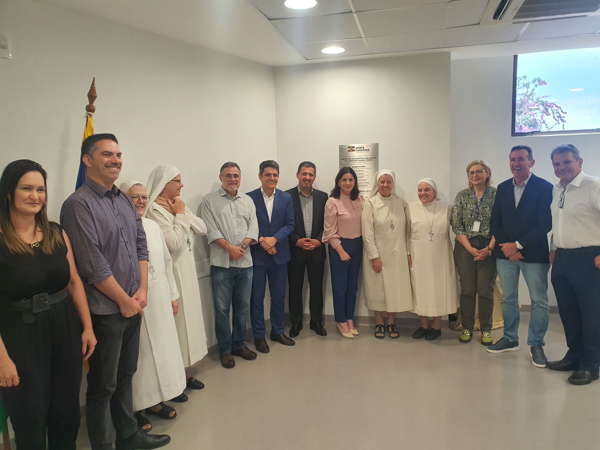 A imagem mostra um grupo de pessoas posando para uma foto em um ambiente interno bem iluminado, possivelmente durante um evento institucional ou inauguração.  Entre os presentes, há homens, mulheres e freiras vestindo hábitos brancos, todos alinhados lado a lado, sorrindo para a câmera. Ao fundo, vê-se uma placa na parede (provavelmente comemorativa ou informativa) e uma bandeira parcialmente visível à esquerda. O clima é formal, mas descontraído, indicando um momento de celebração ou registro oficial.