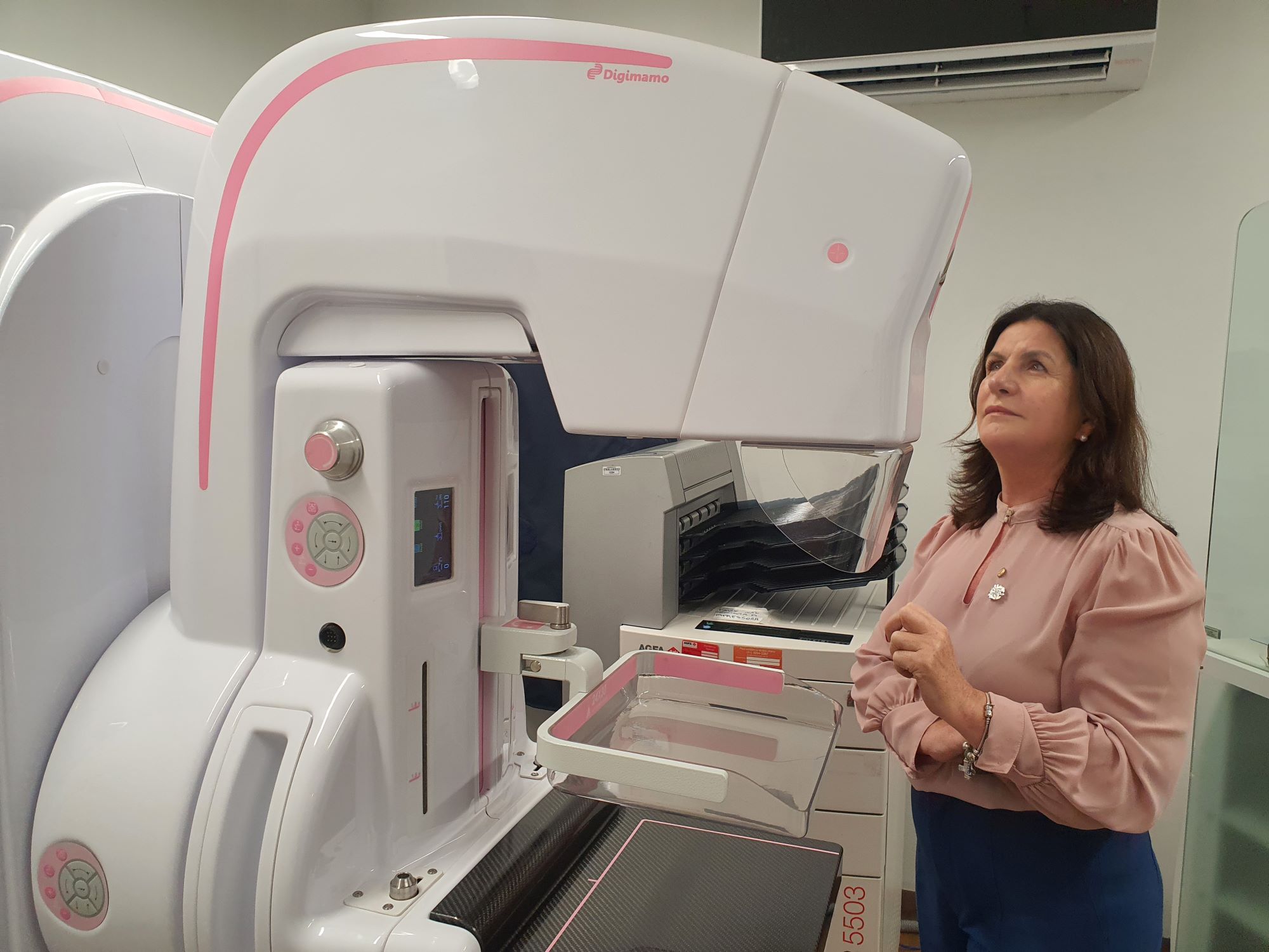 A imagem mostra uma mulher de cabelos castanhos escuros, vestindo blusa rosa clara e calça azul, observando atentamente um equipamento médico de mamografia da marca Giotto Class (DigiMamo), de cor branca com detalhes em rosa. O aparelho é utilizado para exames de detecção e diagnóstico precoce do câncer de mama. Ao fundo, há um aparelho de ar-condicionado instalado na parede e parte de outros equipamentos médicos e mobiliário. O ambiente é limpo e organizado, típico de uma clínica ou hospital.