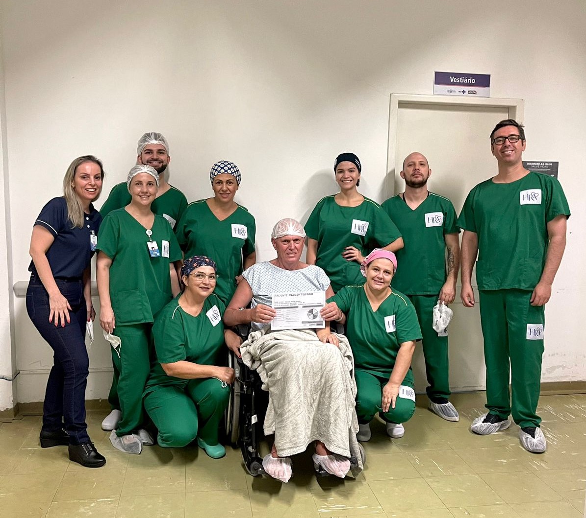 Equipe médica posa ao redor de um paciente em cadeira de rodas, que segura um certificado. Todos usam vestimentas hospitalares, com a maioria dos profissionais vestindo roupas verdes e toucas. A foto é tirada em um corredor de hospital, com uma porta ao fundo identificada como 'Vestiário'.