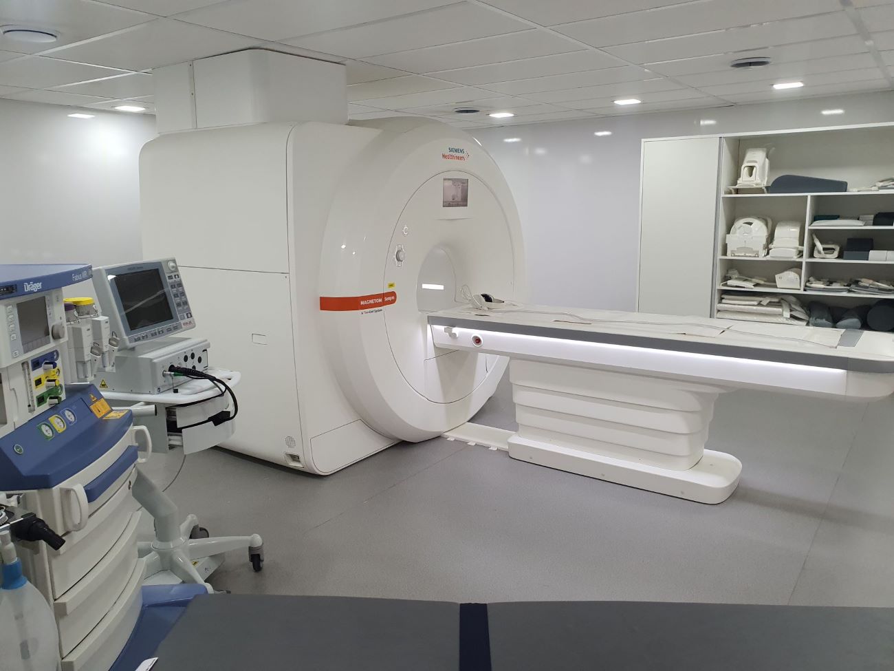 A imagem mostra uma sala hospitalar equipada com um aparelho moderno de ressonância magnética da marca Siemens, modelo Magnetom Sempra. À esquerda, há um equipamento de anestesia/monitoramento da marca Dräger, com telas e controles. No fundo, há prateleiras organizadas com acessórios, bobinas e almofadas utilizadas durante exames de imagem. O ambiente é limpo, bem iluminado e projetado para procedimentos de diagnóstico por imagem.