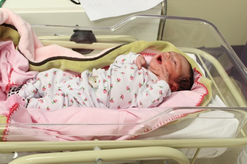 A imagem mostra um recém-nascido deitado em um berço hospitalar transparente, forrado com cobertores cor-de-rosa e verde. O bebê veste um macacão branco estampado com pequenos desenhos coloridos e está com os olhos fechados e a boca aberta, possivelmente chorando. O cenário ao fundo é típico de um ambiente hospitalar, com parte de uma bancada e papéis visíveis.