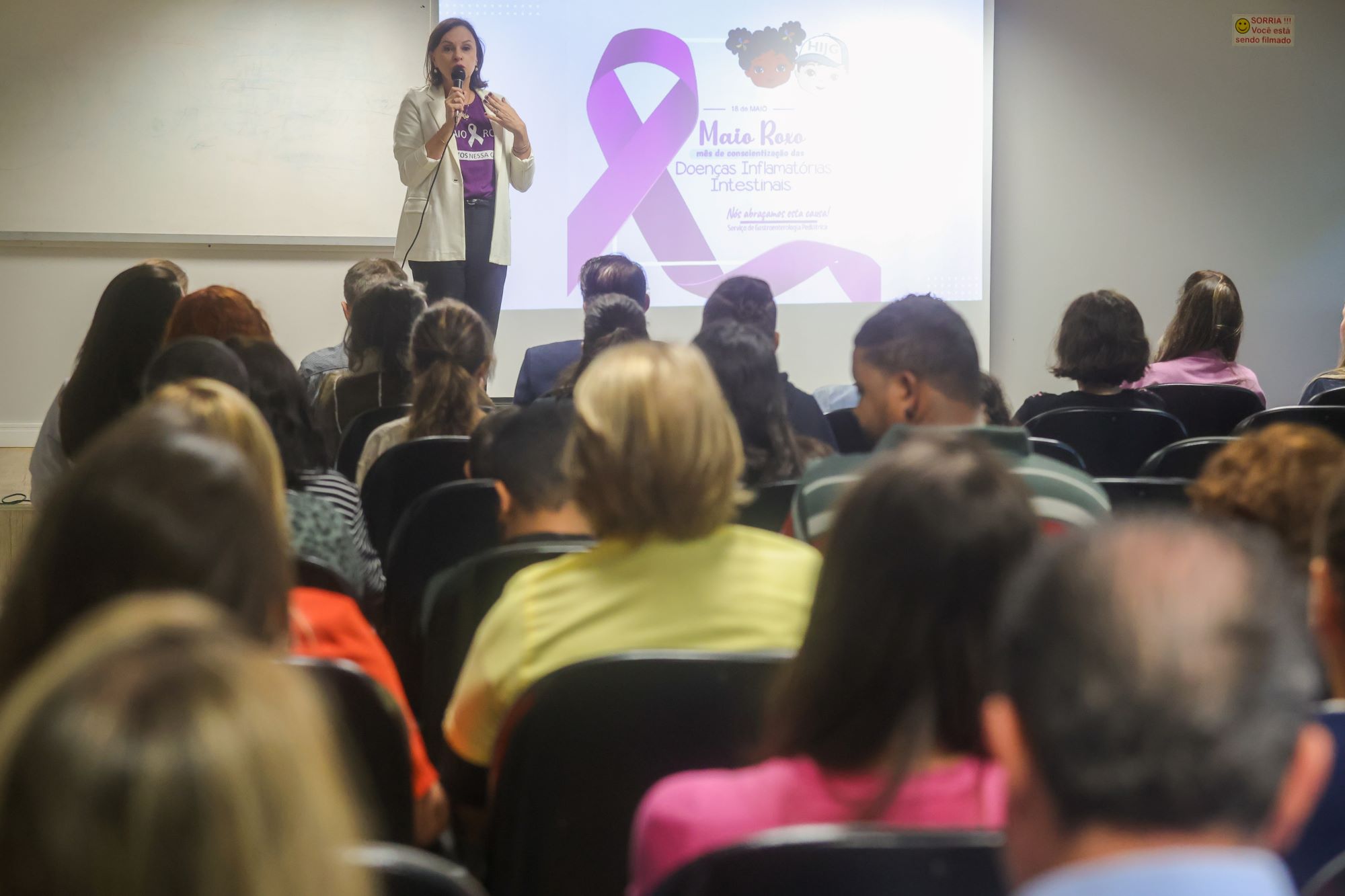 Palestrante fala ao microfone em auditório durante evento do Maio Roxo, mês de conscientização sobre Doenças Inflamatórias Intestinais. Ao fundo, projeção com laço roxo e personagens infantis, diante de uma plateia atenta.