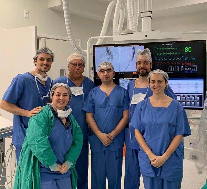 Equipe médica com seis profissionais usando roupas cirúrgicas, toucas e máscaras no pescoço, posando para foto em uma sala cirúrgica. Ao fundo, há monitores com imagens de exames e sinais vitais do paciente. Ambiente esterilizado e equipamentos médicos visíveis.