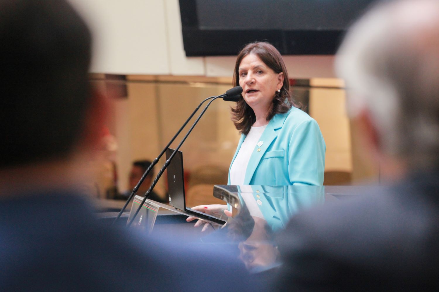 Mulher em pé discursando em um púlpito com microfones duplos, usando blazer azul-claro e blusa branca. Ela está em um ambiente fechado, provavelmente em uma sessão oficial ou evento institucional. Pessoas são vistas desfocadas em primeiro plano.