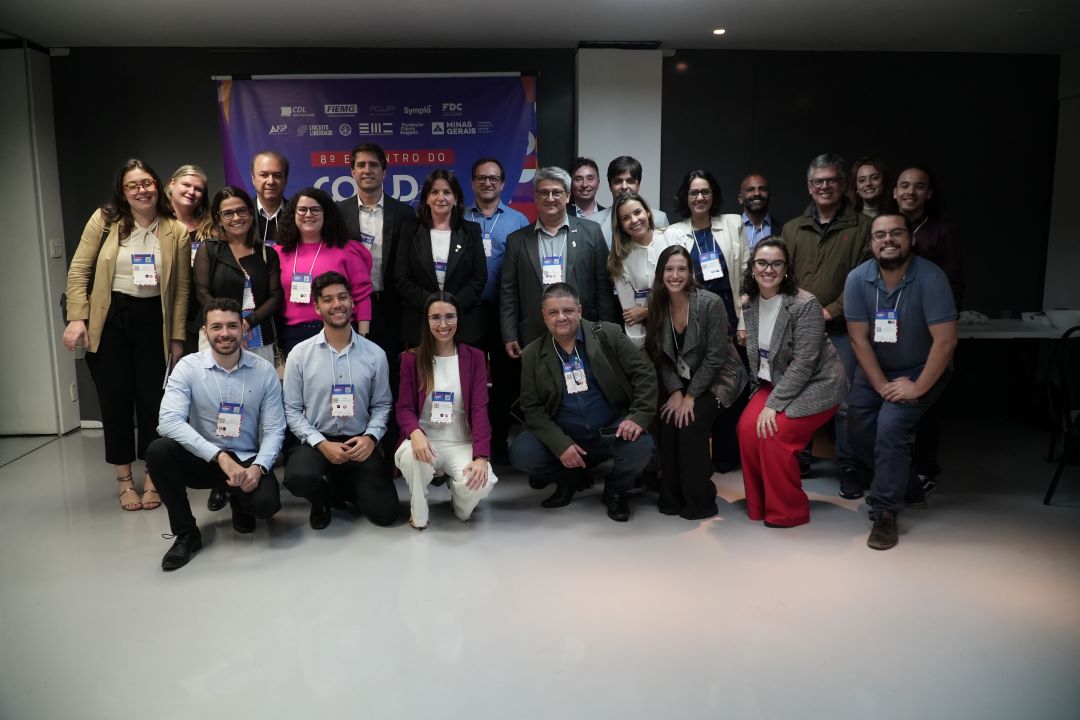 Grupo de cerca de 25 pessoas posando para uma foto em um evento profissional, diante de um banner com logotipos de entidades como CDL, FIEMG e FDC. Os participantes, usando crachás, estão sorrindo, alguns agachados na frente e outros em pé ao fundo, demonstrando um momento de confraternização e networking.