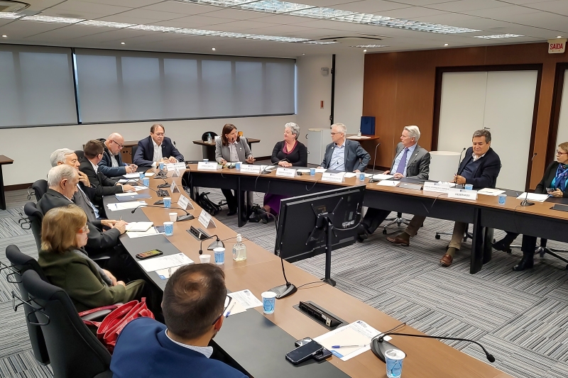 Grupo de pessoas reunidas em torno de uma mesa retangular em uma sala de reuniões corporativa. Todos estão sentados, em trajes formais, participando de uma discussão. Há nomeações sobre a mesa, copos de café e microfones, indicando um encontro formal ou reunião institucional.