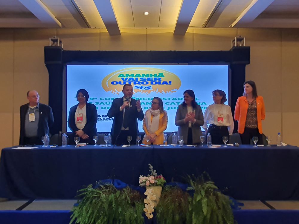 Autoridades de saúde em pé atrás de uma mesa durante a 9ª Conferência Estadual de Saúde de Santa Catarina. Um homem ao centro fala ao microfone, cercado por mulheres e homens com crachás de identificação. Ao fundo, um telão exibe o tema do evento: 'Amanhã Vai Ser Outro Dia! SUS'. A mesa está coberta por uma toalha azul e decorada com arranjos de samambaias e flores.