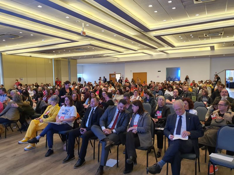 Auditório lotado com dezenas de pessoas sentadas assistindo à 9ª Conferência Estadual de Saúde de Santa Catarina. Na frente, autoridades e participantes com crachás estão atentos, alguns conversando entre si. O ambiente é amplo, bem iluminado, com teto rebaixado e painéis de luz.