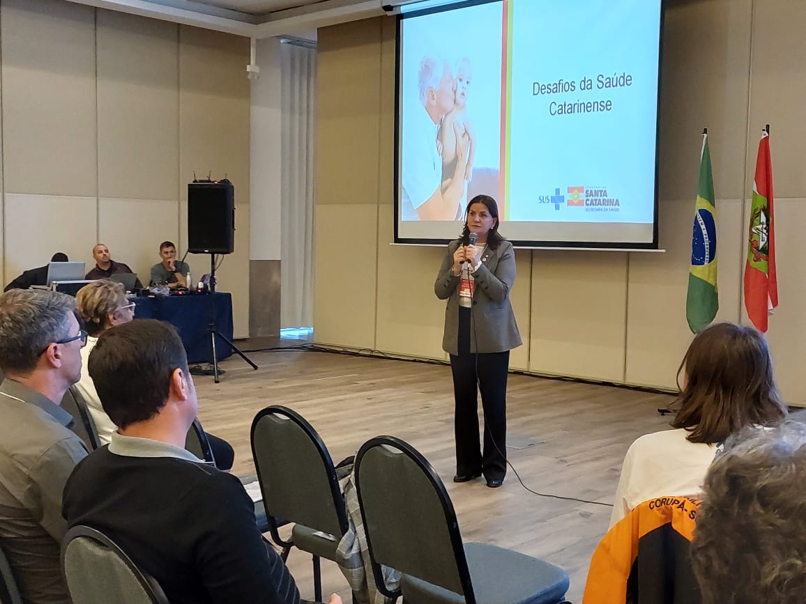 Secretária de Estado da Saúde de Santa Catarina discursa em pé com microfone em mãos, diante de uma plateia, durante a 9ª Conferência Estadual de Saúde. Ao fundo, um telão exibe a apresentação com o título 'Desafios da Saúde Catarinense' e logotipos do SUS e do Governo de Santa Catarina. À direita do telão, estão as bandeiras do Brasil e de Santa Catarina. Participantes assistem atentos, sentados.