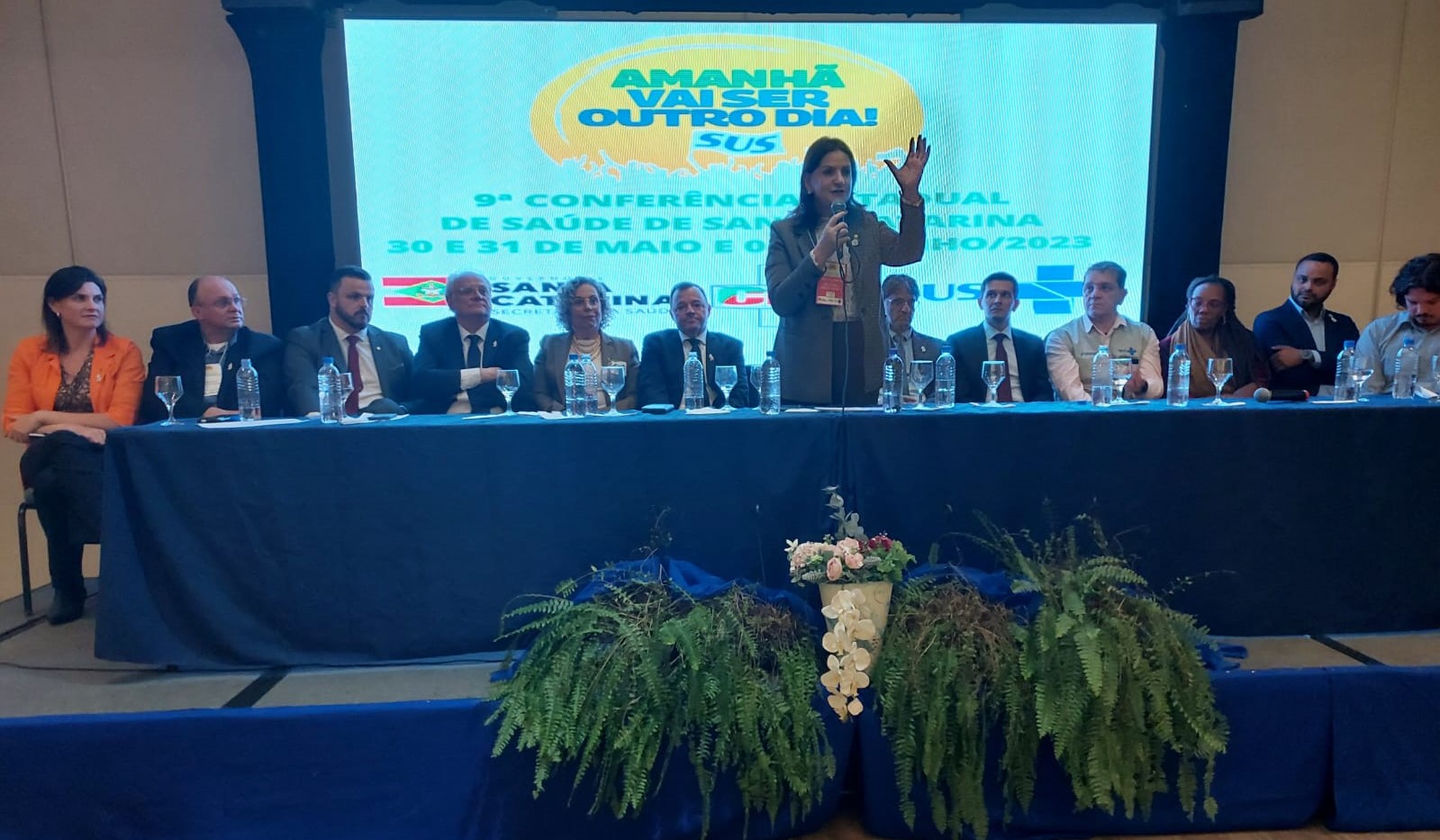 Autoridades e representantes da saúde participam da 9ª Conferência Estadual de Saúde de Santa Catarina. Uma mulher, em pé ao centro, discursa com microfone diante de uma mesa composta por diversas pessoas sentadas. Ao fundo, o telão exibe o tema do evento: ‘Amanhã vai ser outro dia! SUS’, com datas de realização: 30 e 31 de maio e 1º de junho de 2023.