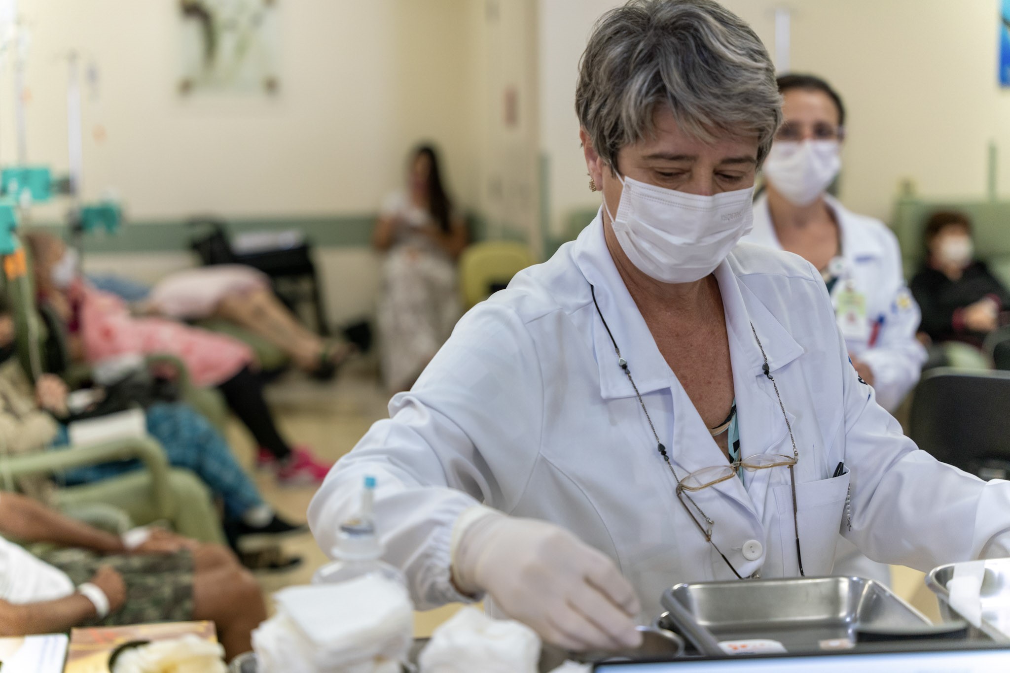 Profissional de saúde em ambiente hospitalar, usando máscara e luvas, manuseia materiais médicos em uma bandeja. Ao fundo, outros profissionais e pacientes estão sentados em poltronas, indicando um possível ambiente de quimioterapia ou enfermaria especializada.