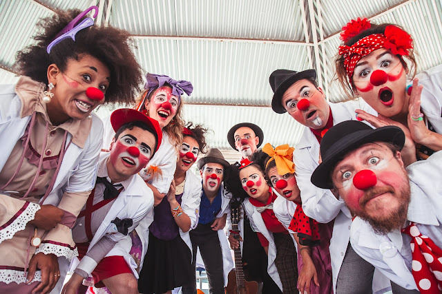 Grupo de palhaços reunido sob uma estrutura metálica, todos com narizes vermelhos e expressões animadas e surpresas, olhando para a câmera. Vestem roupas coloridas e criativas, com jalecos brancos, chapéus, laços e adereços variados. A cena transmite alegria e descontração.