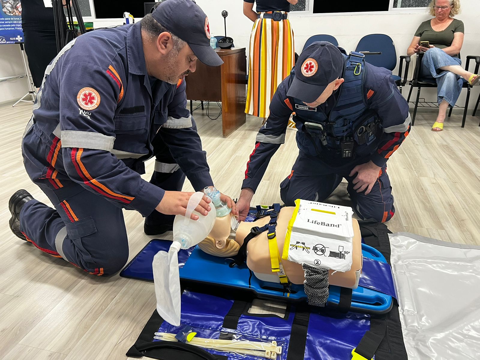 A imagem mostra dois profissionais do SAMU, vestidos com uniformes azul-marinho e faixas refletivas, realizando um treinamento de atendimento de emergência.  Eles estão ajoelhados no chão e utilizam um manequim para simular uma vítima, aplicando técnicas de ressuscitação cardiopulmonar (RCP) com auxílio de um equipamento de ventilação manual (Ambu) e um dispositivo de compressão torácica automática LifeBand.  Ao fundo, há pessoas observando, indicando que se trata de uma atividade instrutiva ou de capacitação.