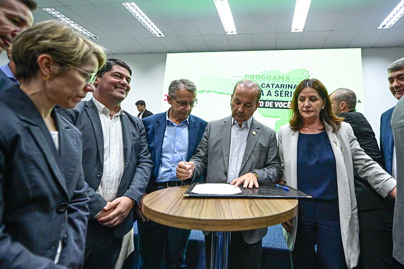 Grupo de autoridades em um evento institucional, com um homem ao centro assinando um documento sobre uma mesa redonda. Ao fundo, tela digital com texto "Programa Santa Catarina Leva a Sério - Perto de Você". Todos estão com trajes formais e focados na cerimônia.