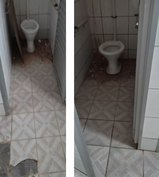 A imagem mostra dois boxes de banheiro pequenos, ambos com vasos sanitários do tipo sem caixa acoplada, instalados junto à parede de fundo.  As paredes são revestidas com azulejos brancos e o piso é de cerâmica clara com padrão geométrico em tons de bege. No box da esquerda, há entulho e sujeira acumulados no chão, especialmente perto do vaso, além de uma placa ou pedaço de material encostado na lateral. O box da direita também apresenta sujeira, mas em menor quantidade, e sinais de desgaste na base das paredes. Ambos os espaços estão em estado de abandono ou pré-reforma, necessitando de limpeza e manutenção.