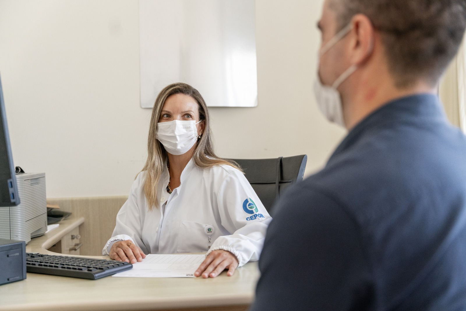 Profissional de saúde do CEPON, usando jaleco branco e máscara, atende paciente mascarado em consultório médico, com mesa, computador e papéis à frente.