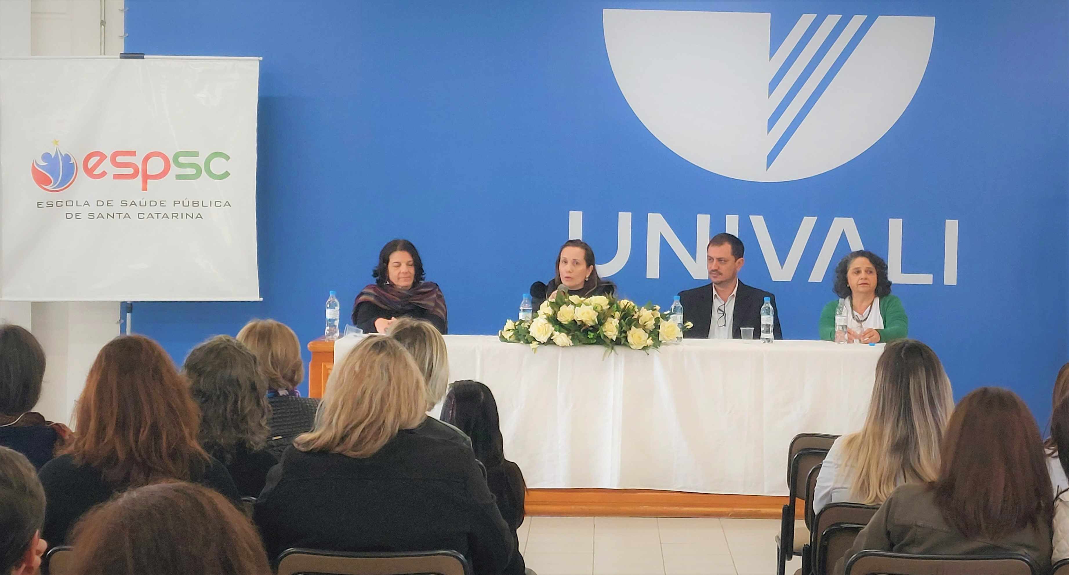 Quatro pessoas sentadas em uma mesa de abertura em frente ao público em auditório da UNIVALI. À esquerda, banner da ESP-SC (Escola de Saúde Pública de Santa Catarina). Ao centro da mesa, arranjo de flores brancas e garrafas de água. Fundo azul com logomarca da UNIVALI.